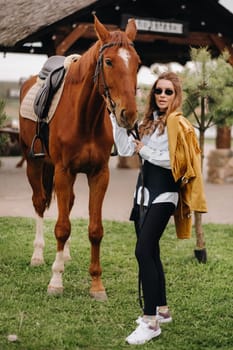 Stylish girl with glasses stands next to a horse on the street.