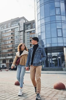 Cheerful couple in casual warm clothes have a walk outdoors in the city near business building.
