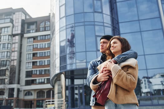 Cheerful couple in casual warm clothes have a walk outdoors in the city near business building.