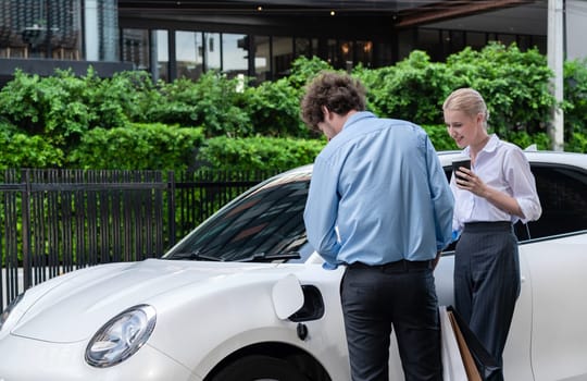 Progressive businessman and businesswoman with electric car parking and connected to public charging station before driving around city center. Eco friendly rechargeable car powered by clean energy.