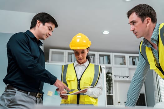 A team of competent engineers wearing safety equipment is working on blueprints with a tablet while also discussing with the investor in the office.