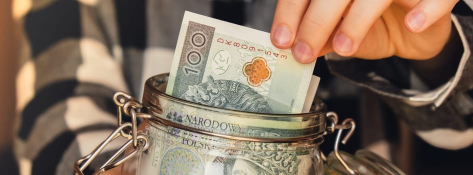 Polish zloty banknote saving money in glass jar. Unrecognizable woman moderate consumption and economy Collecting money. Tips. Business, finance, saving, banking and people concept. Extra money, passive income. Saving for travel, crisis, house, car, education, retirement Banner