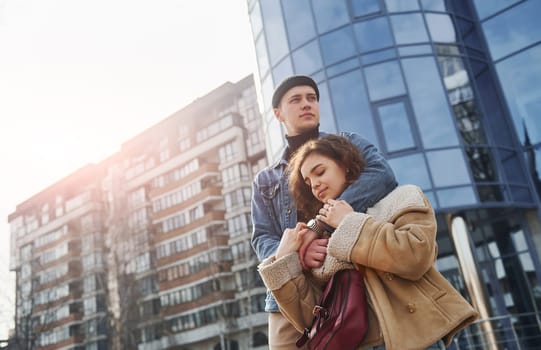 Cheerful couple in casual warm clothes have a walk outdoors in the city near business building.