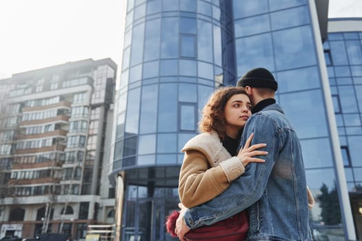 Cheerful couple in casual warm clothes have a walk outdoors in the city near business building.