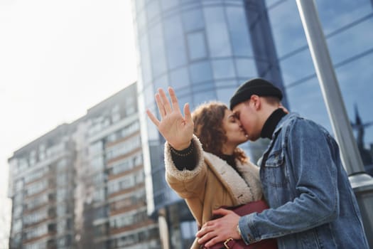 Cheerful couple in casual warm clothes kissing outdoors in the city near business building.