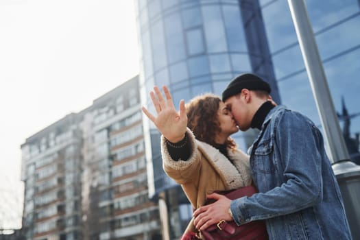 Cheerful couple in casual warm clothes kissing outdoors in the city near business building.