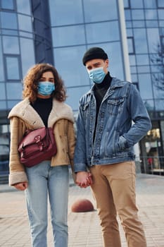 Couple in protective masks have a walk outdoors in the city near business building at quarantine time. Conception of coronavirus.