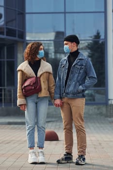 Couple in protective masks have a walk outdoors in the city near business building at quarantine time. Conception of coronavirus.