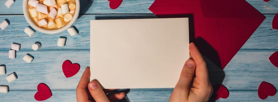 Banner Female hands holding empty valentine card mock up red envelope with white cup of coffee and marshmallows on wooden blue background. Romantic Small hearts Valentine day. Blank paper card copy space for your text. Holiday morning. Top view, flat lay minimalist branding concept