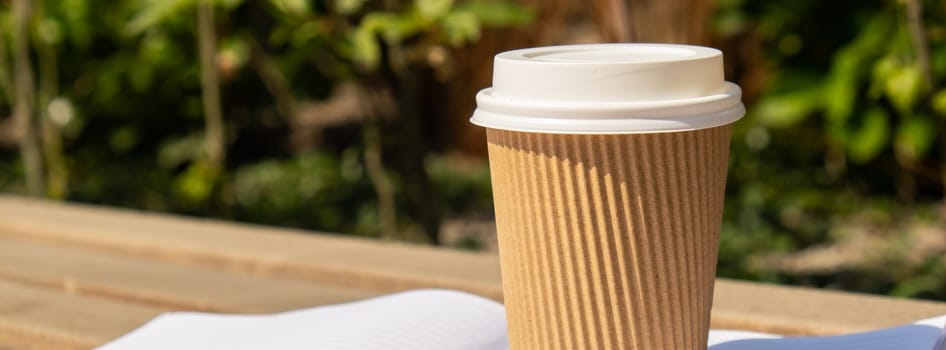 Eco recycling paper cup with coffee or tea on kraft paper with empty paper notebook on wooden bench. Concept of study work outdoors. Take away coffee to go. Copy space for text. Disposable Cardboard coffee outdoors
