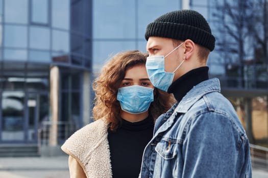 Couple in protective masks have a walk outdoors in the city near business building at quarantine time. Conception of coronavirus.