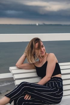 Young woman sitting on bench on wooden pier blurred beachside background. Attractive female enjoying the sea shore travel and active lifestyle concept. Springtime. Wellness wellbeing mental health inner peace Slow life digital detox