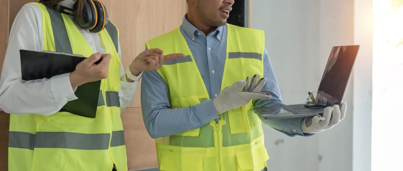 engineer construction and in vest with helmet working with laptop, standing on building construction site. Home building project. Engineer discusses with coworker at workplace.