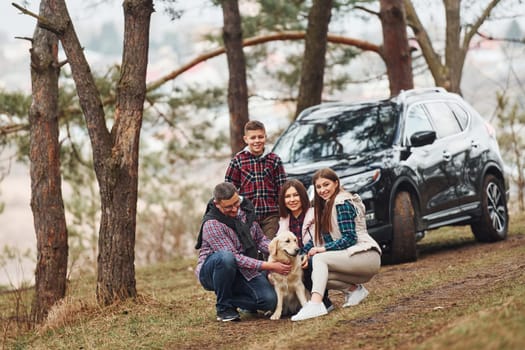 Happy family sitting and having fun with their dog near modern car outdoors in forest.