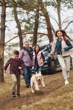 Happy family runs and having fun with their dog near modern car outdoors in forest.