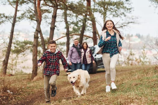 Happy family runs and having fun with their dog near modern car outdoors in forest.