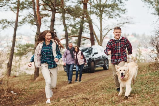 Happy family runs and having fun with their dog near modern car outdoors in forest.