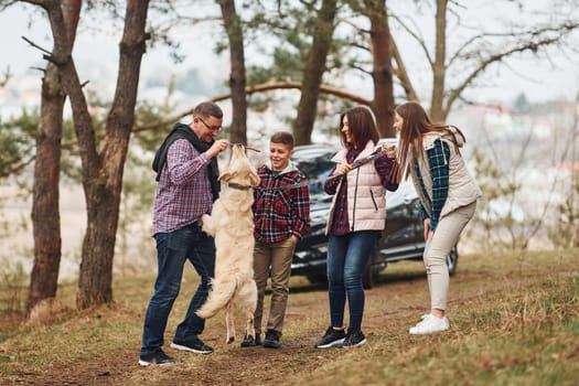 Happy family have fun with their active dog near modern car outdoors in forest.