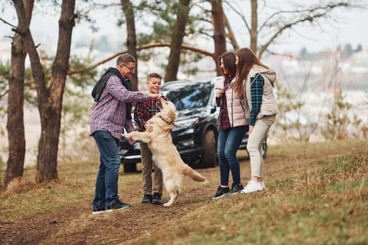 Happy family have fun with their active dog near modern car outdoors in forest.