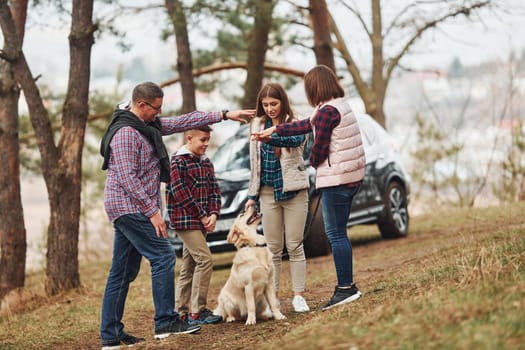 Happy family have fun with their active dog near modern car outdoors in forest.