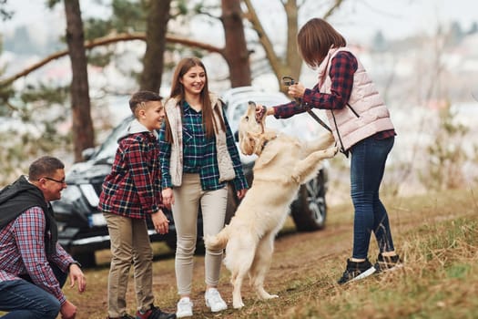 Happy family have fun with their active dog near modern car outdoors in forest.