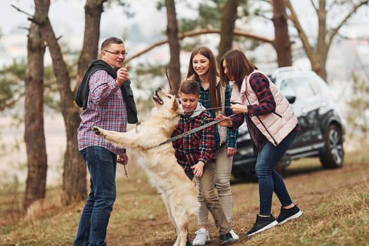 Happy family have fun with their active dog near modern car outdoors in forest.