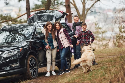 Happy family have fun with their dog near modern car outdoors in forest.