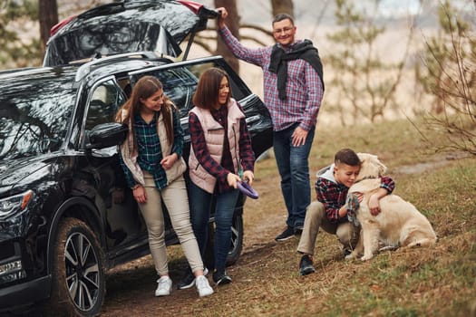 Happy family have fun with their dog near modern car outdoors in forest.