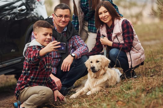 Happy family sitting and having fun with their dog near modern car outdoors in forest.