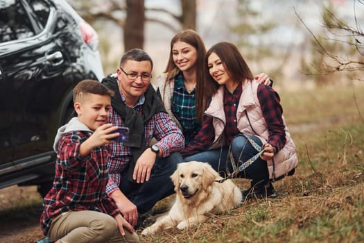 Happy family sitting and having fun with their dog near modern car outdoors in forest.