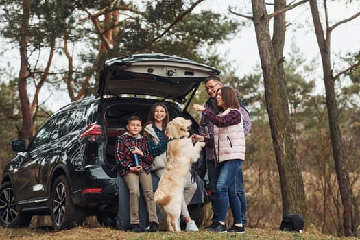 Happy family have fun with their dog near modern car outdoors in forest.