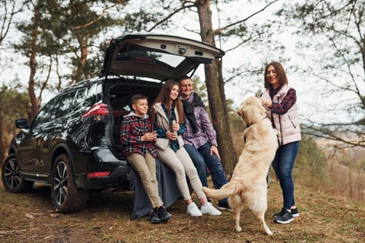 Happy family have fun with their dog near modern car outdoors in forest.