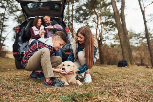 Happy family sitting and having fun with their dog near modern car outdoors in forest.