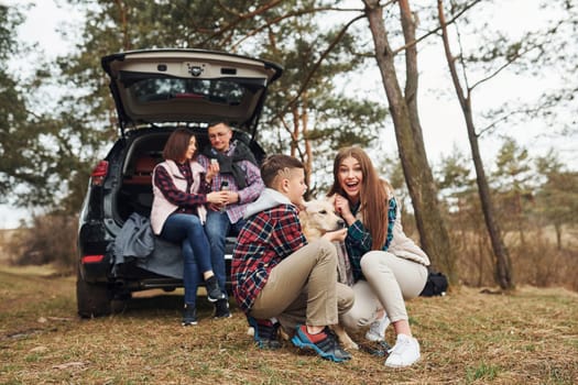 Happy family sitting and having fun with their dog near modern car outdoors in forest.