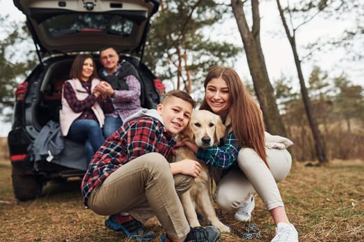 Happy family sitting and having fun with their dog near modern car outdoors in forest.