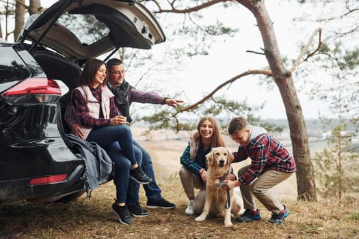 Happy family sitting and having fun with their dog near modern car outdoors in forest.