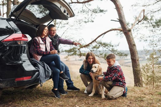 Happy family sitting and having fun with their dog near modern car outdoors in forest.