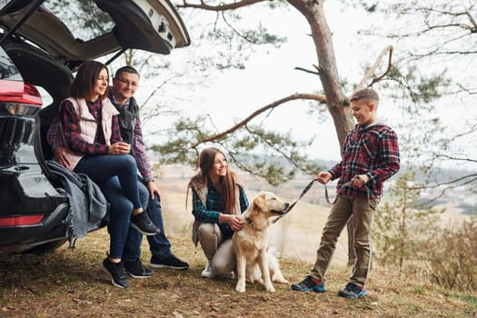 Happy family sitting and having fun with their dog near modern car outdoors in forest.