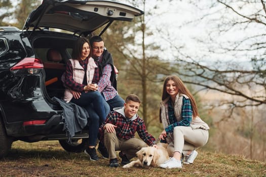 Happy family sitting and having fun with their dog near modern car outdoors in forest.