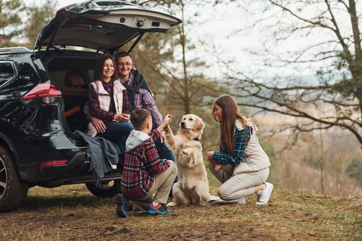 Happy family have fun with their dog near modern car outdoors in forest.