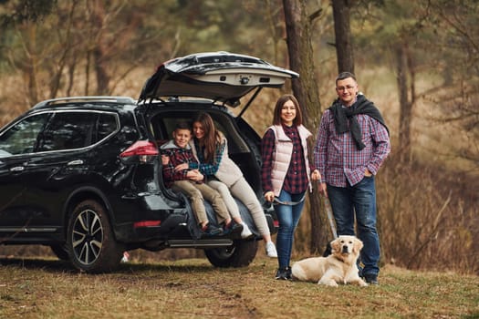 Happy family have fun with their dog near modern car outdoors in forest.