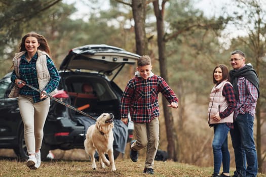 Sister and brother runs forward. Happy family have fun with their dog near modern car outdoors in forest.