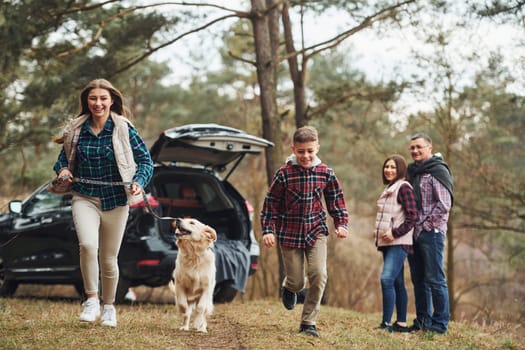 Sister and brother runs forward. Happy family have fun with their dog near modern car outdoors in forest.