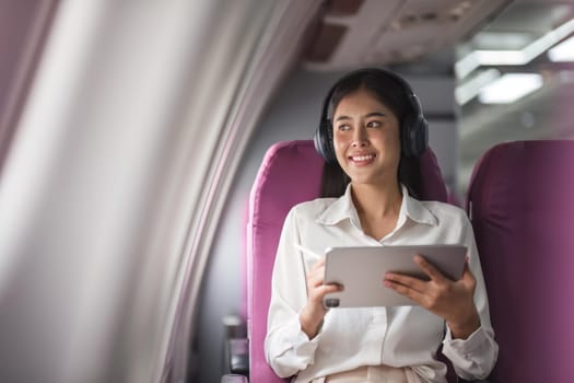 Cheerful female passenger in headphones for noise cancellation watching online movie during intercontinental flight in cabin of aircraft, happy young woman using wifi connection on board.