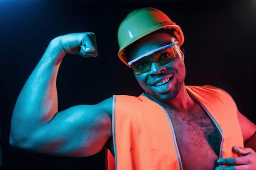 Construction worker in uniform and hard hat. Futuristic neon lighting. Young african american man in the studio.