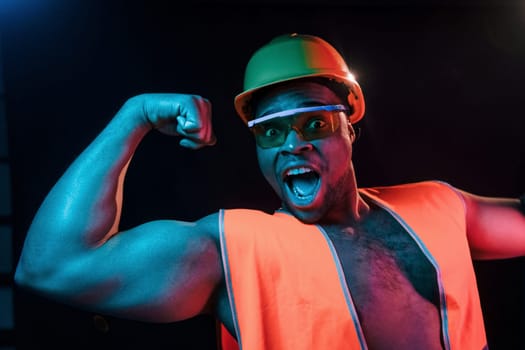 Construction worker in uniform and hard hat. Futuristic neon lighting. Young african american man in the studio.