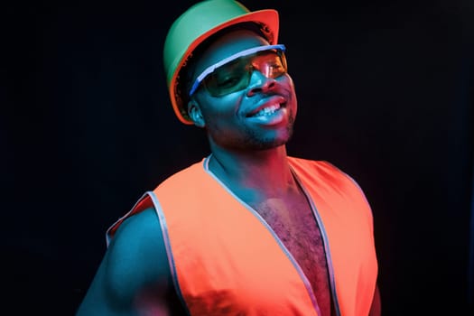 Construction worker in uniform and hard hat. Futuristic neon lighting. Young african american man in the studio.