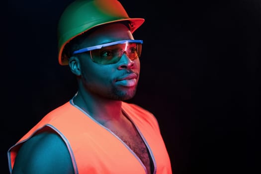 Construction worker in uniform and hard hat. Futuristic neon lighting. Young african american man in the studio.