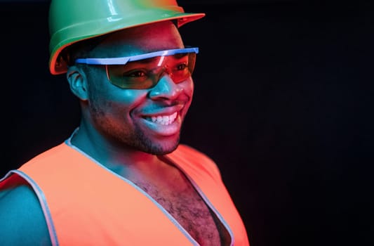 Construction worker in uniform and hard hat. Futuristic neon lighting. Young african american man in the studio.