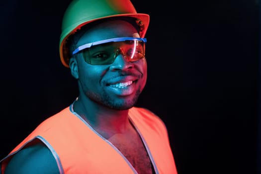 Construction worker in uniform and hard hat. Futuristic neon lighting. Young african american man in the studio.